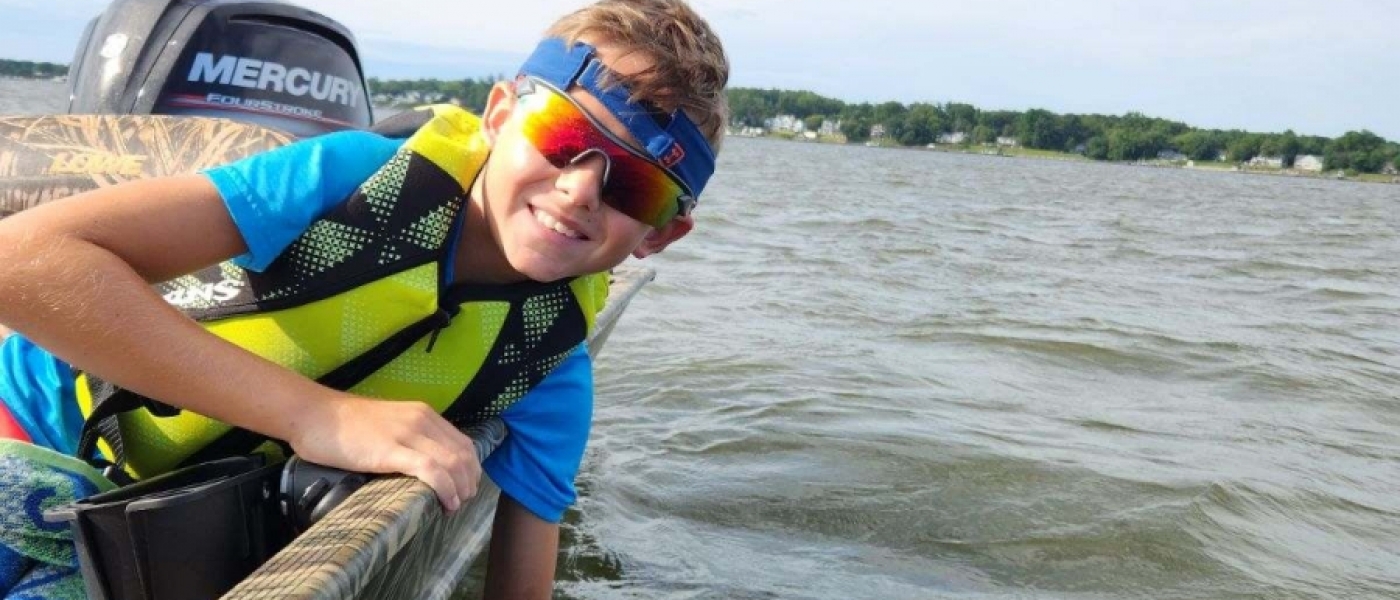 A young boy in sunglasses and a neon yellow life vest leans over the side of a boat, holding a water bottle