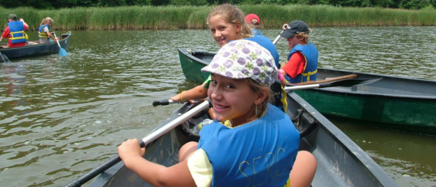Two young girls in blue life vests padding a canoe, turning around to smile at the camera