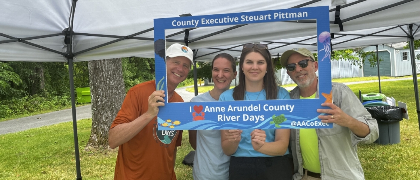 Four people pose while holding a blue frame with the words "County Executive Steuart Pittman. Anne Arundel County River Days"