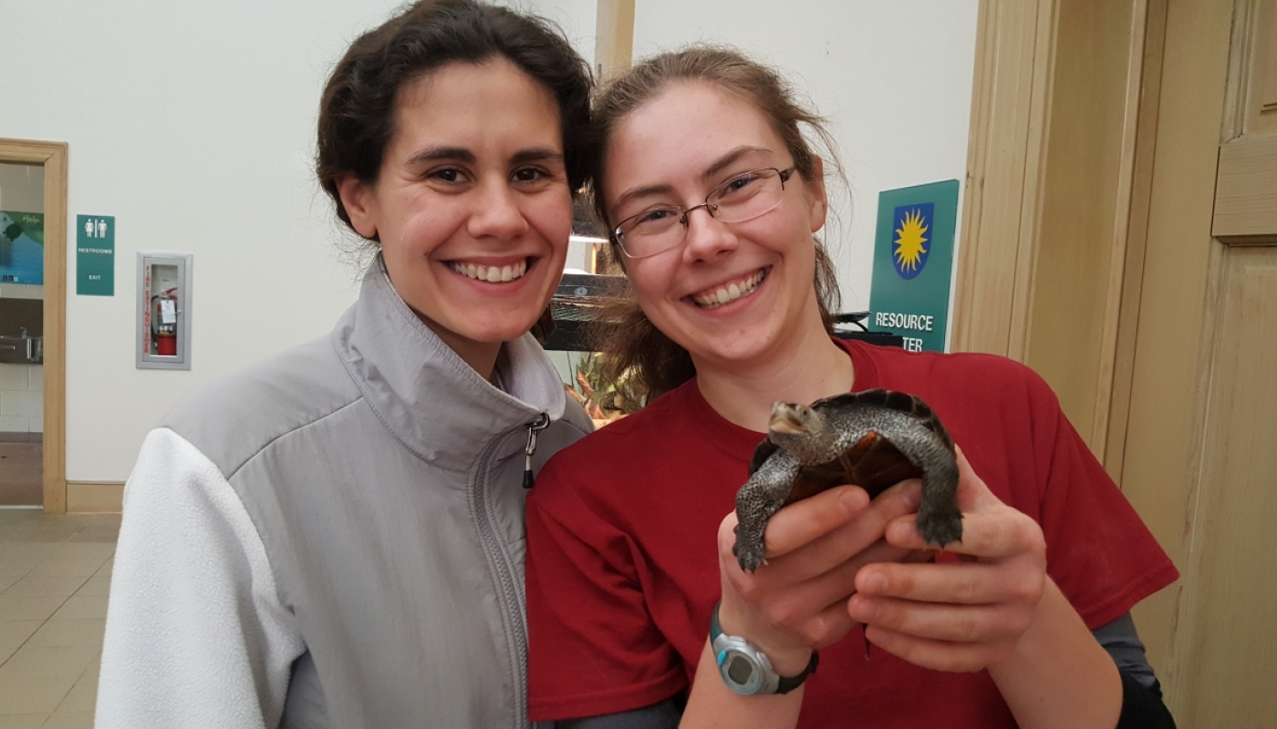 Two young women stand side by side, smiling. The one on the right holds a turtle in her hands