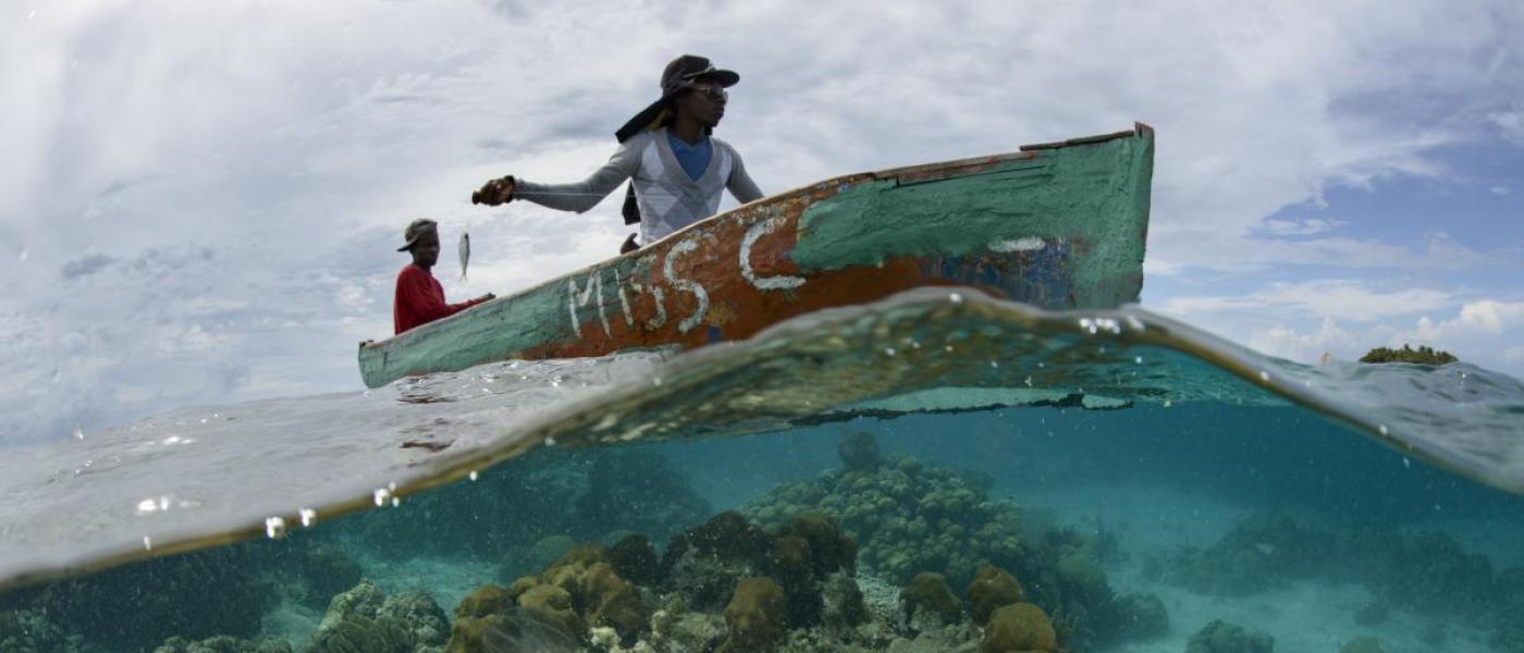 Two dark-skinned people in a small boat, one holding a small fish on a line. Bottom of the photo shows underwater view of reef