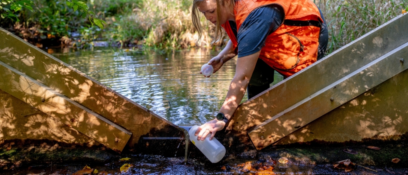 A woman in a bright orange vest kneeling to collect water from a weir into a cylindrical container