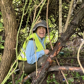 Research Technician, Jessie Ribera, smiles at camera through small opening in thorn patch, while squatting below vegetation.