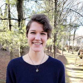 A smiling white woman with short asymmetrical hair, dark blue sweater, and necklace against a background of trees.