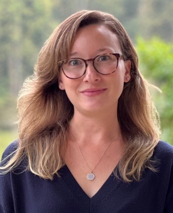 A headshot of Anna Davis, who has shoulder-length blond hair, glasses, and is wearing a blue sweater, in front of a forest.