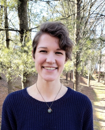 A smiling white woman with short asymmetrical hair, dark blue sweater, and necklace against a background of trees.