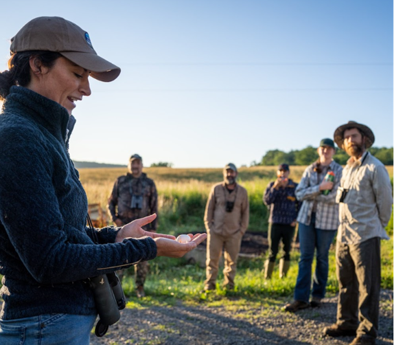 A woman--Dr. Amy Johnson--wearing a baseball cap, navy blue sweatshirt, and jeans, holding a small item in her hand and talking about it, while 5 people are gathered around, listening to her. They are in an open field under a light blue sky.